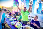 A man in a bright green shirt stands and raises a water bottle joyfully at an outdoor event in the Big Easy, while seated people around a table look on.