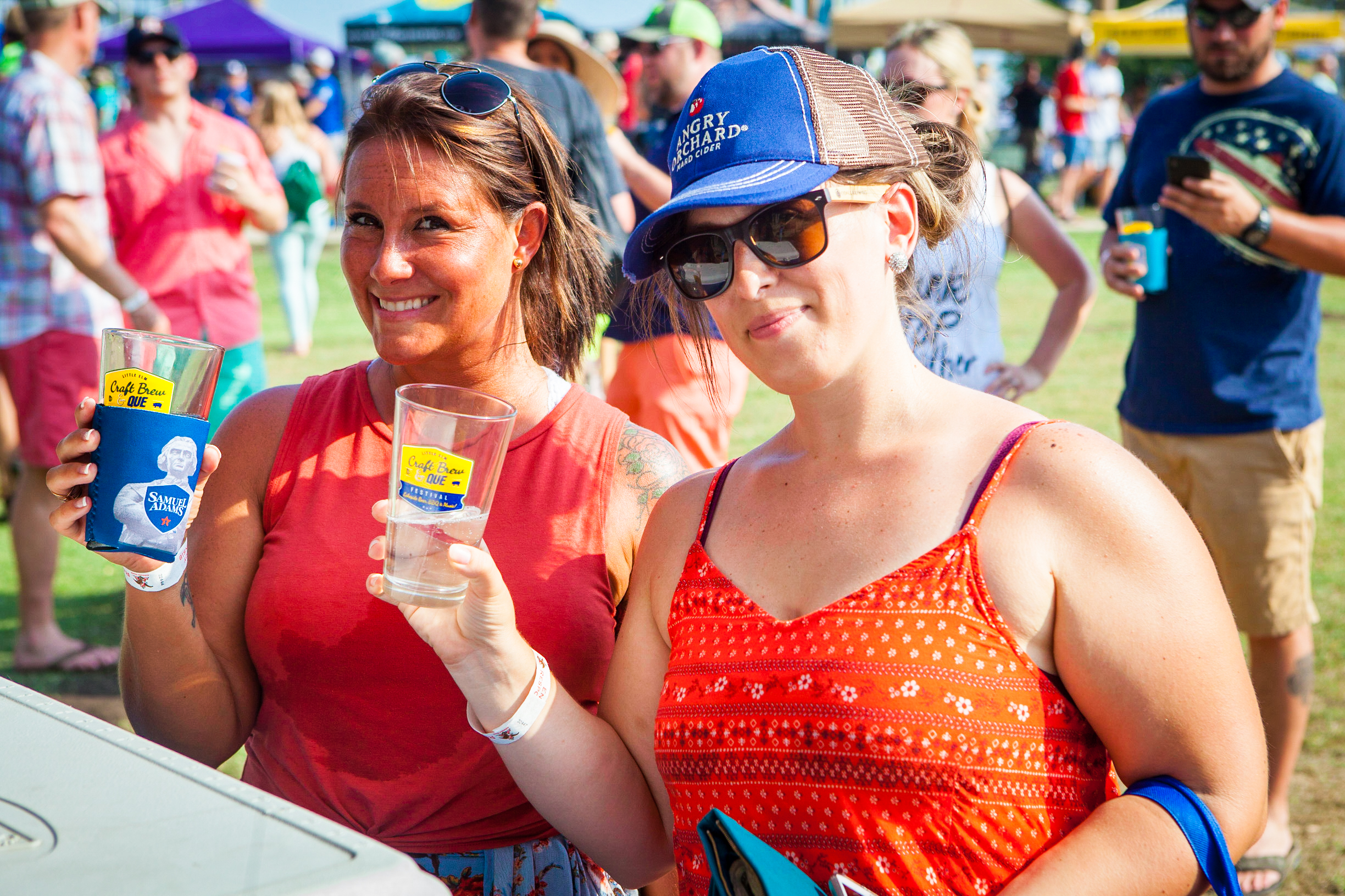 Two women holding clear plastic cups of beer enjoy Brew & Que festivities, surrounded by a lively crowd and colorful tents at the outdoor event.