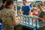 A person in a camouflage shirt pours beer from a row of taps at the outdoor Brew & Que event, where people, including a curious child, are eagerly waiting and observing.