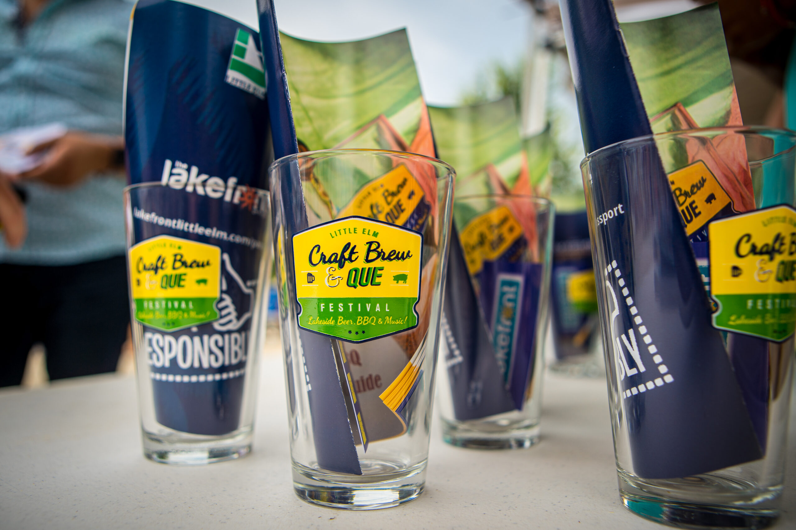 Several Brew & Que festival branded pint glasses sit on the table, each containing pamphlets celebrating the art of craft beer and BBQ.