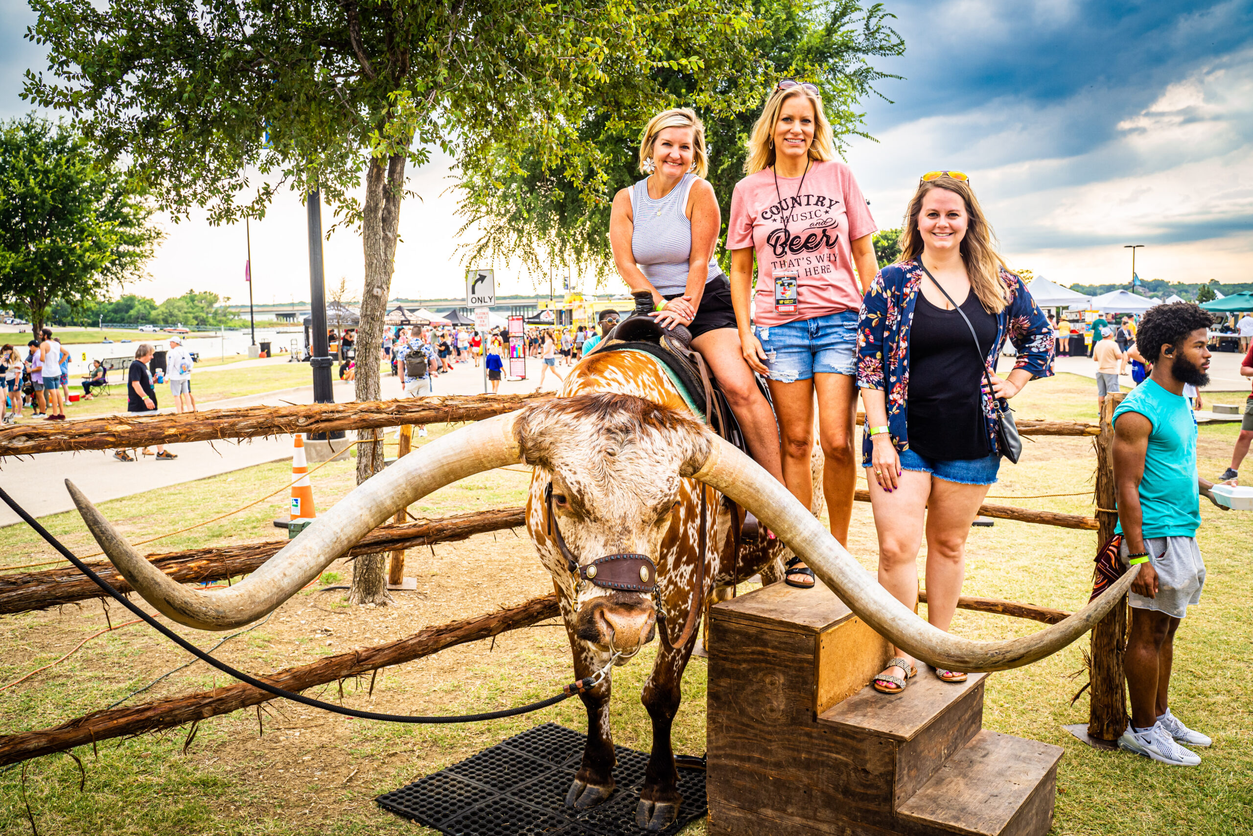 Three women pose on a platform with a longhorn steer in a fenced area at the Brew & Que event, where the aroma of BBQ and freshly poured beer fills the air.