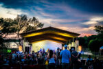 Enjoy a relaxing outdoor concert at the park pavilion during sunset, with a crowd seated on the grass and a stage lit up. Savor local Brew & Que delights with barbeque flavors and refreshing beer, as trees sway gently by the river in the background.
