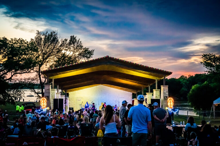 Enjoy a relaxing outdoor concert at the park pavilion during sunset, with a crowd seated on the grass and a stage lit up. Savor local Brew & Que delights with barbeque flavors and refreshing beer, as trees sway gently by the river in the background.