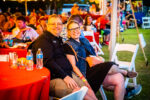 A smiling man and woman sit together at a red table outdoors during the Brew & Que event. With the tempting aroma of BBQ in the air, other attendees can be seen enjoying their beers in the background.