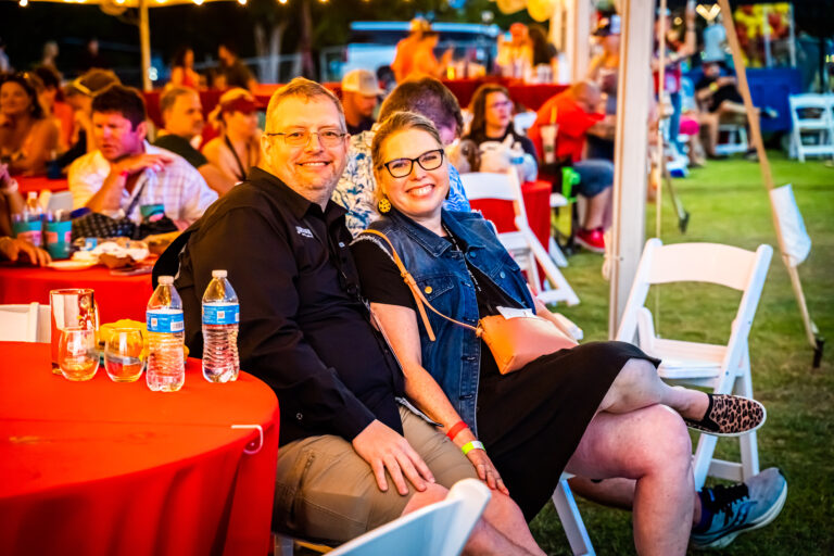 A smiling man and woman sit together at a red table outdoors during the Brew & Que event. With the tempting aroma of BBQ in the air, other attendees can be seen enjoying their beers in the background.