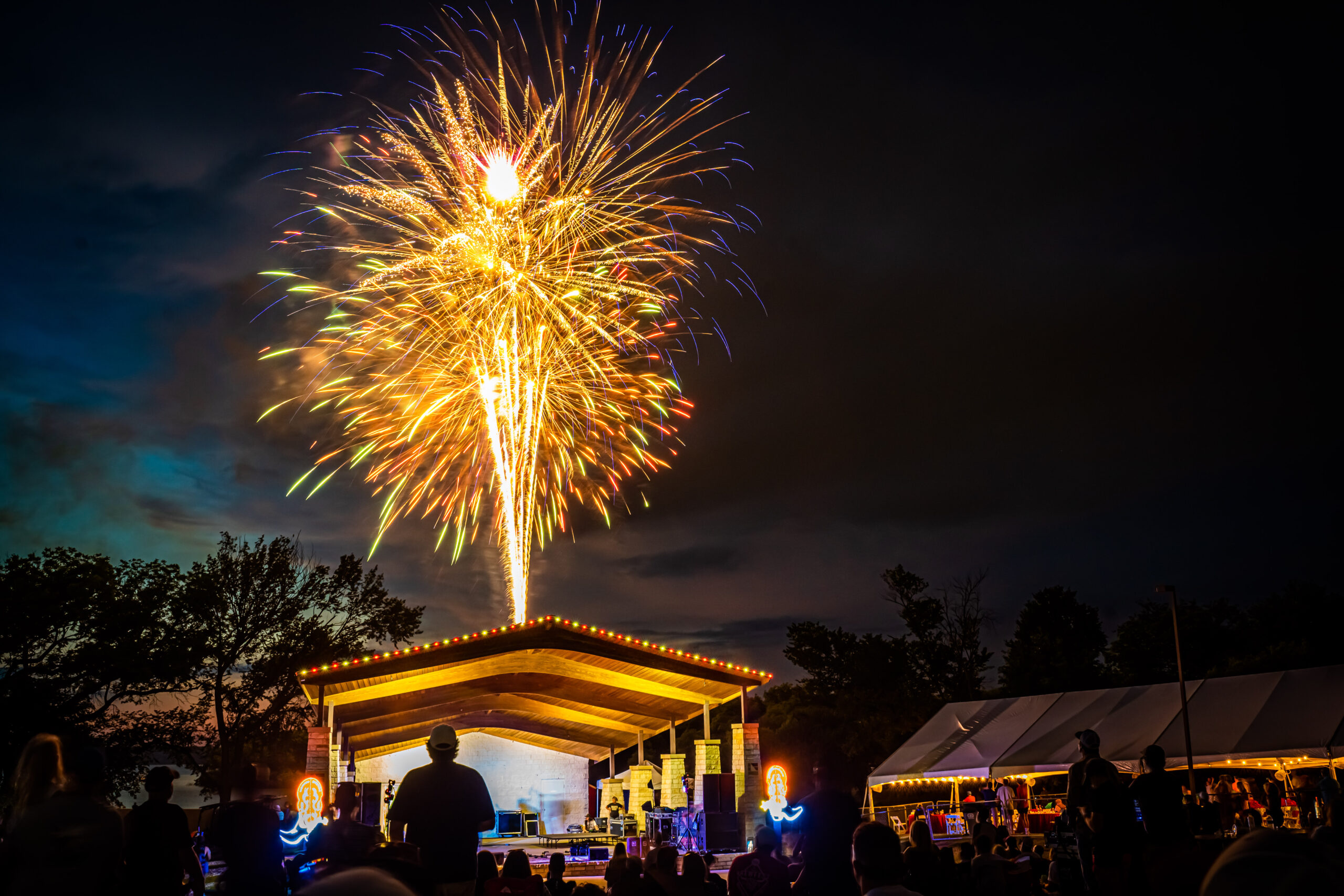 Fireworks burst over an outdoor stage at night, illuminating the scene with colorful light. Silhouetted people watch the display, savoring fresh BBQ and cold beer from Brew & Que, enhancing the vibrant atmosphere.