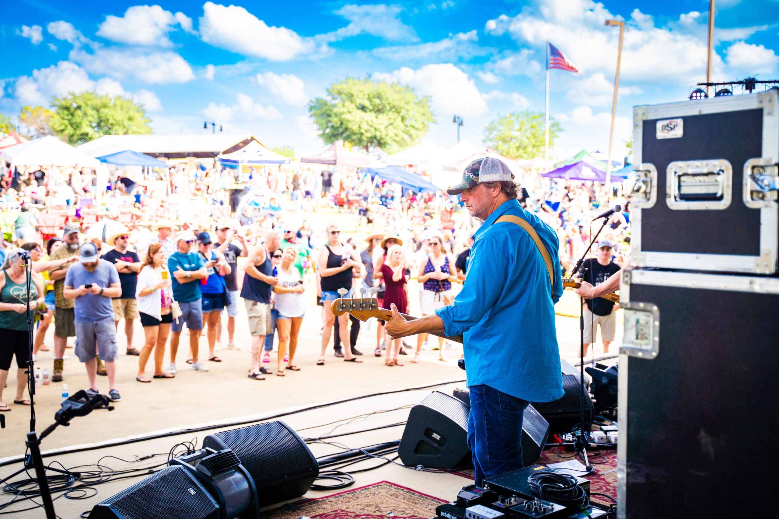 A musician plays guitar on stage at an outdoor Brew & Que festival, with a large crowd enjoying the vibe. Sunny weather and an American flag enhance the scene, while the aroma of BBQ drifts through and cold beer flows freely.