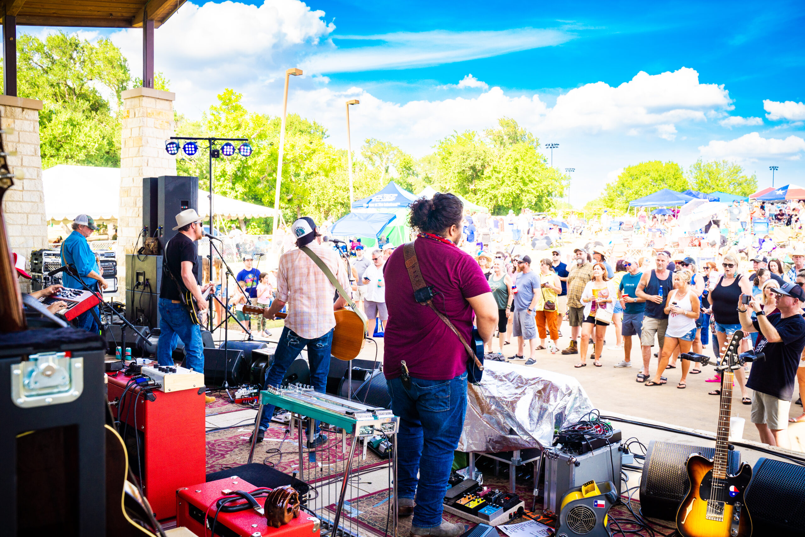 A band performs on an outdoor stage with a crowd gathered in front, enjoying the Brew & Que festival. It's a sunny day with a clear blue sky, perfect for savoring some BBQ and beer.