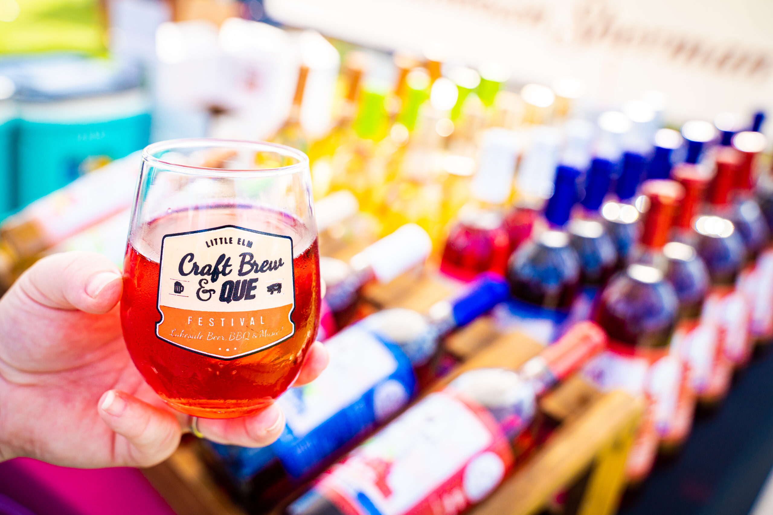 A person holds a glass of craft beer in front of a vibrant display of colorful bottled drinks at the Brew & Que festival.