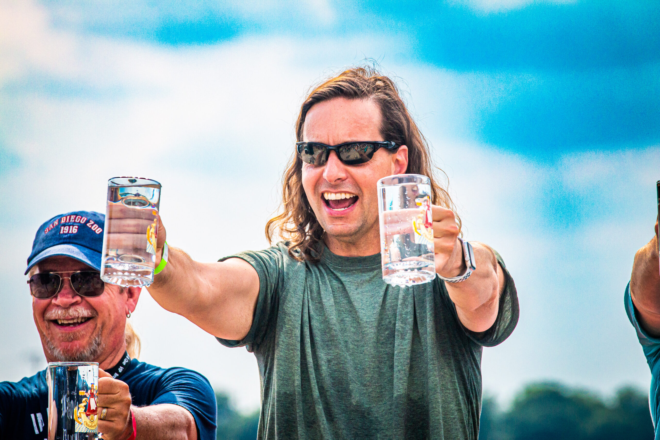 A person with long hair and sunglasses holds a frothy beer mug, smiling outdoors at the Brew & Que event. Another person stands beside them, also savoring their drink. The vibrant blue sky sets the perfect backdrop for this cheerful BBQ gathering.