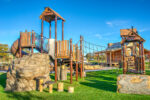 A colorful playground with climbing structures, slides, and wooden elements on a green turf surface, perfect for spring break adventures. Signs read "Kids Only.