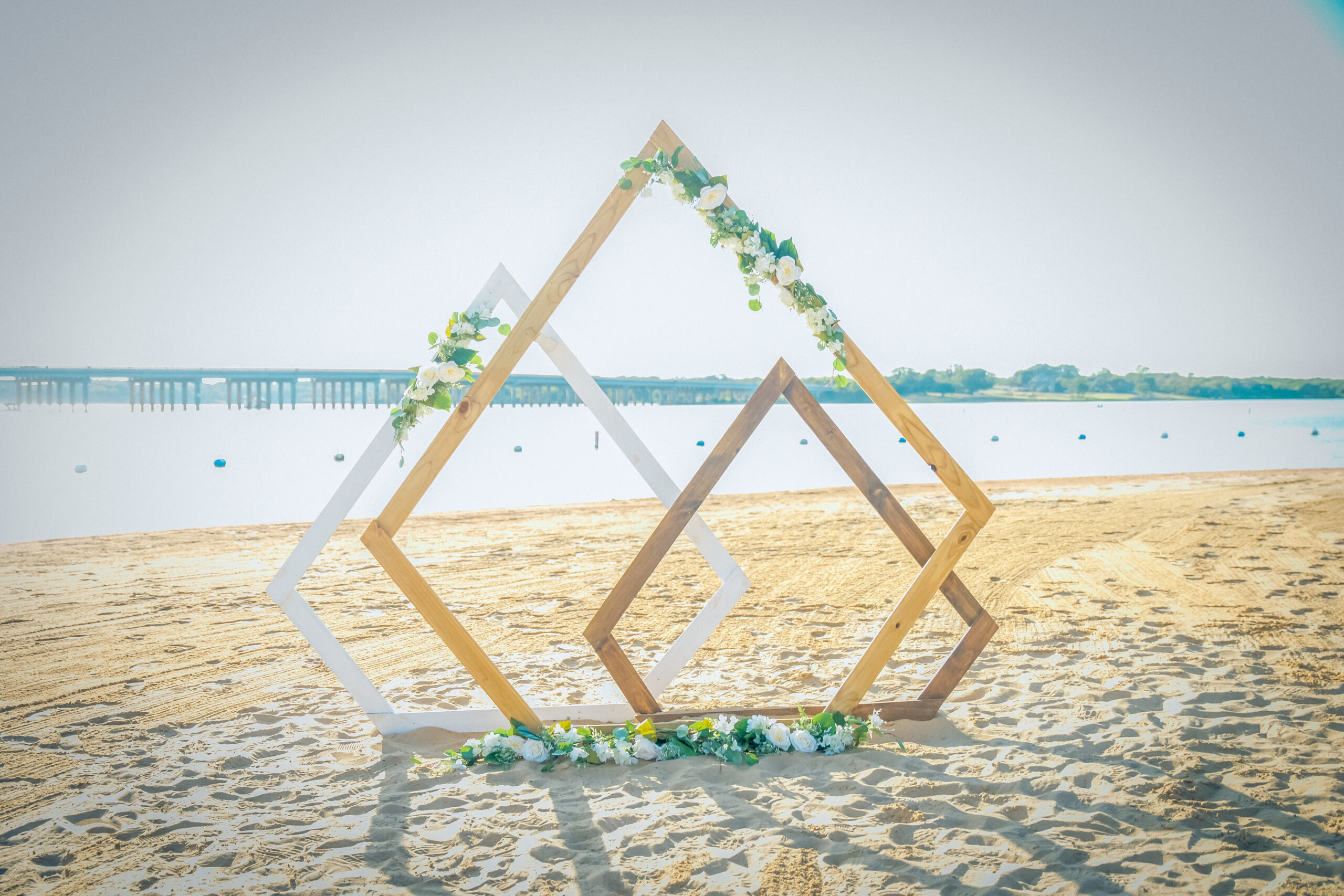 Geometric wooden frames adorned with flowers stand on a sandy beach with the calm waters of Lewisville Lake as a breathtaking backdrop, perfect for beach weddings in Texas.