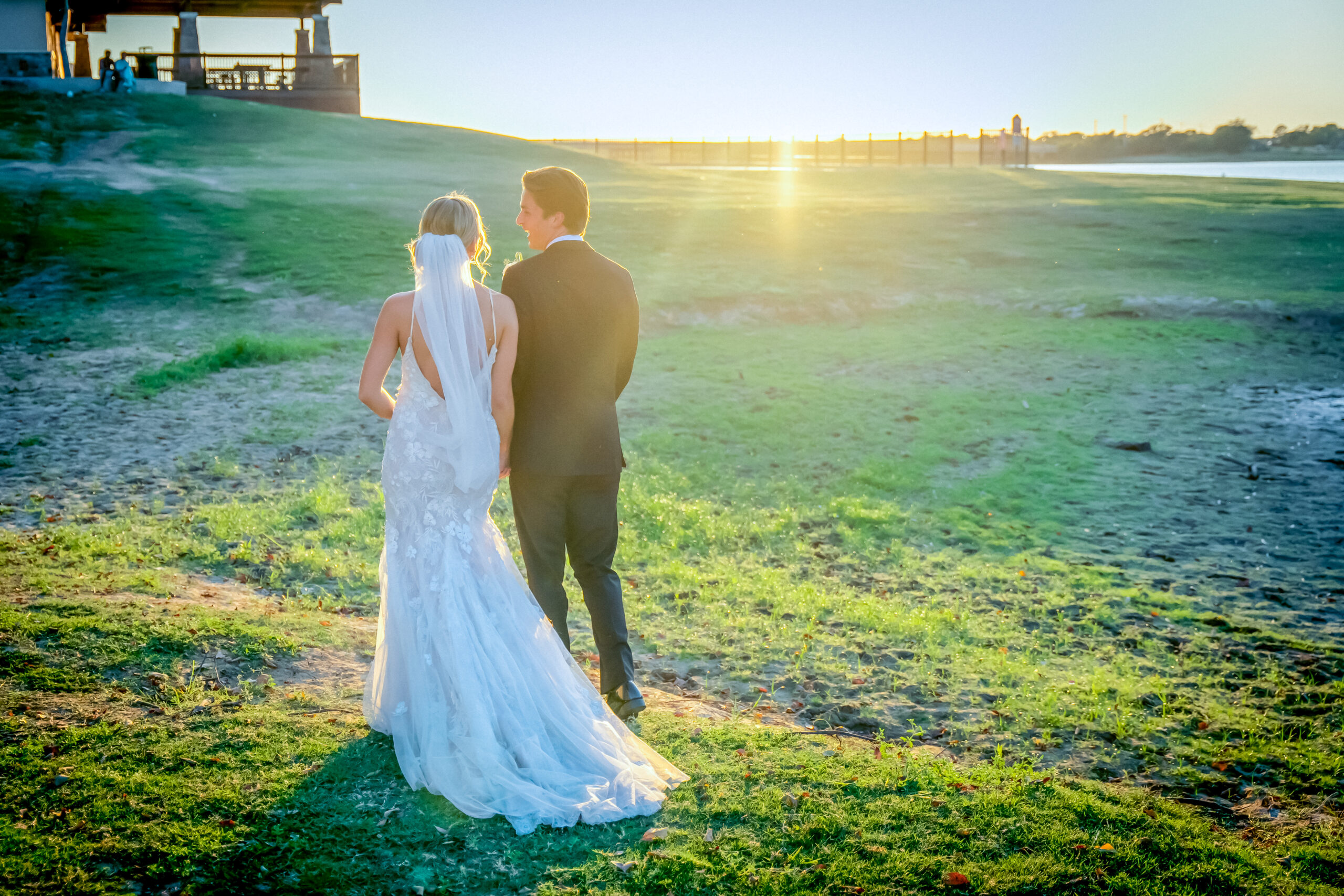 A bride and groom stand outdoors on grass at sunset, the bride in a white gown and veil, the groom in a dark suit, facing a sunlit horizon—perfect for capturing the magic of beach weddings in Texas.