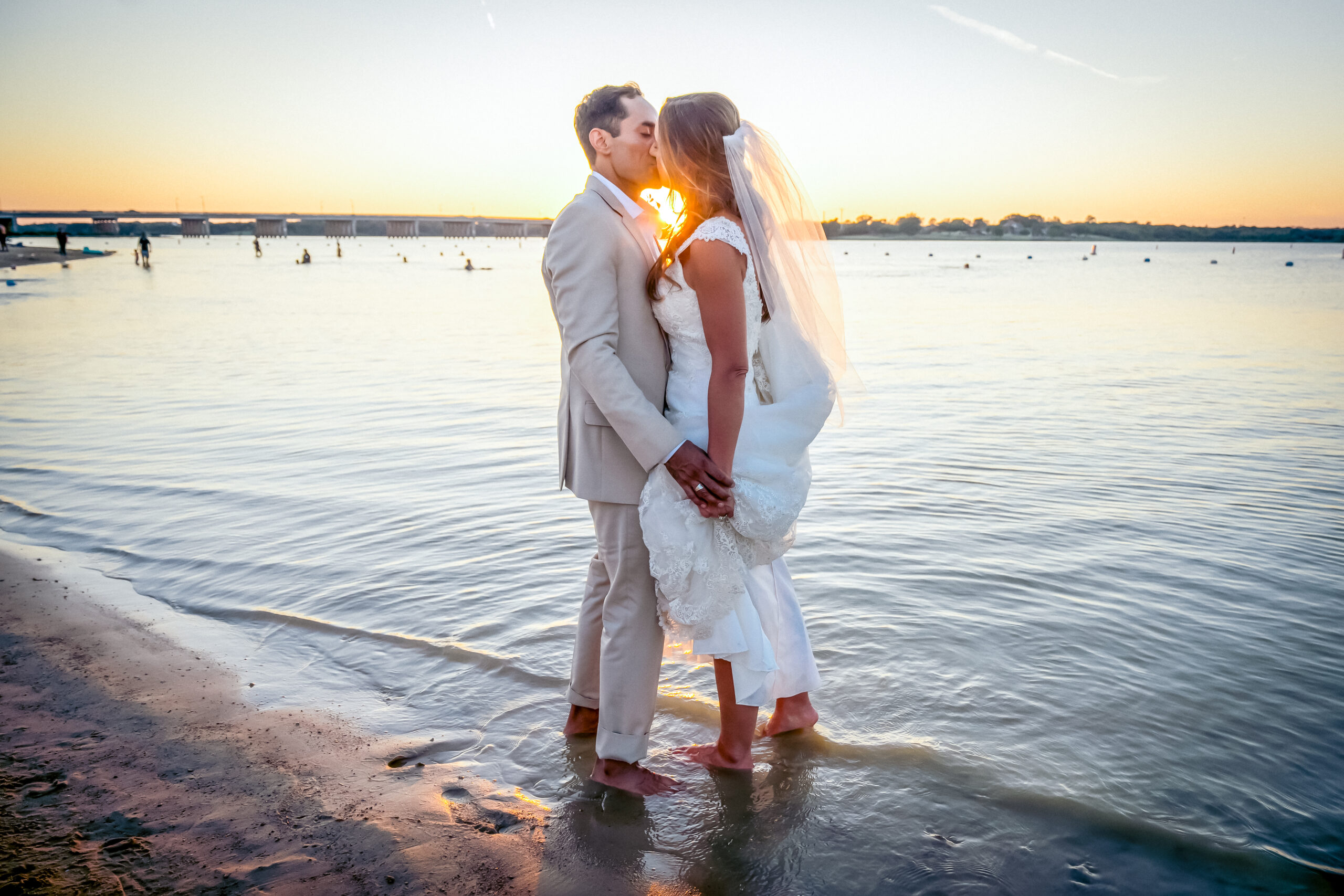 A bride and groom kiss barefoot on a beach at sunset, as gentle waves touch their feet—a magical scene reminiscent of lakeside weddings in Little Elm.
