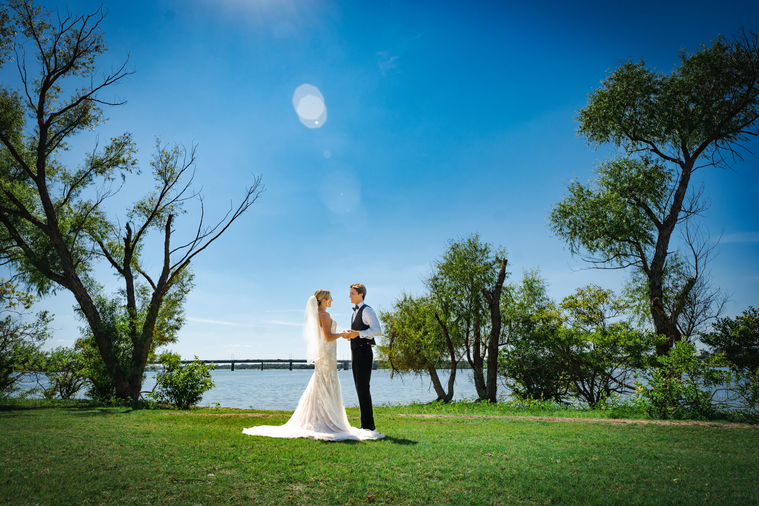 The bride and groom stand facing each other on a grassy area by a lake, surrounded by trees, under a clear blue sky—perfect for Lakeside weddings in Little Elm.
