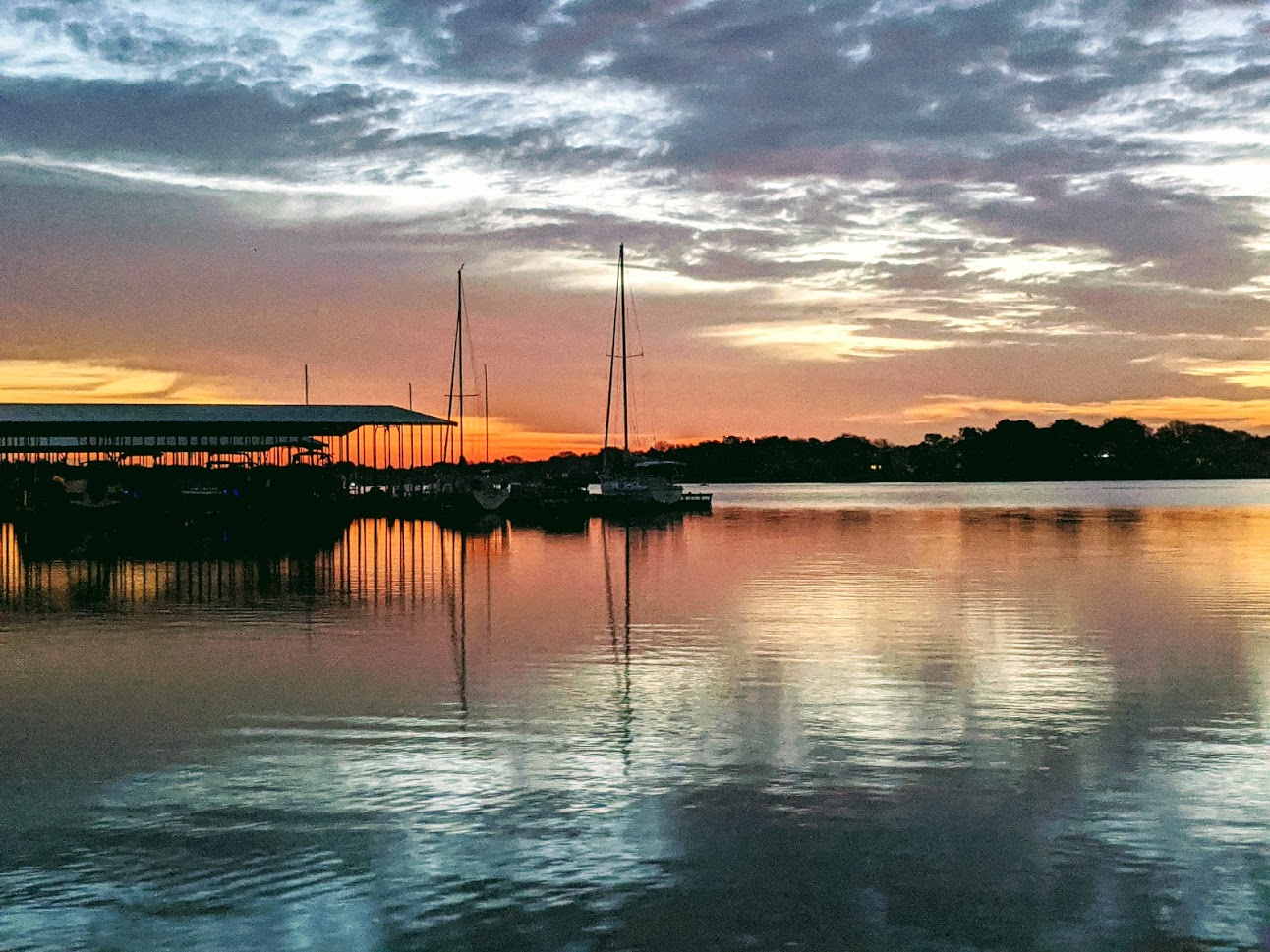 A serene sunset over a calm lake at The Venues at the Lakefront, with sailboats docked at a pier, silhouetted against a colorful sky—a perfect backdrop for unforgettable North Texas wedding venues.