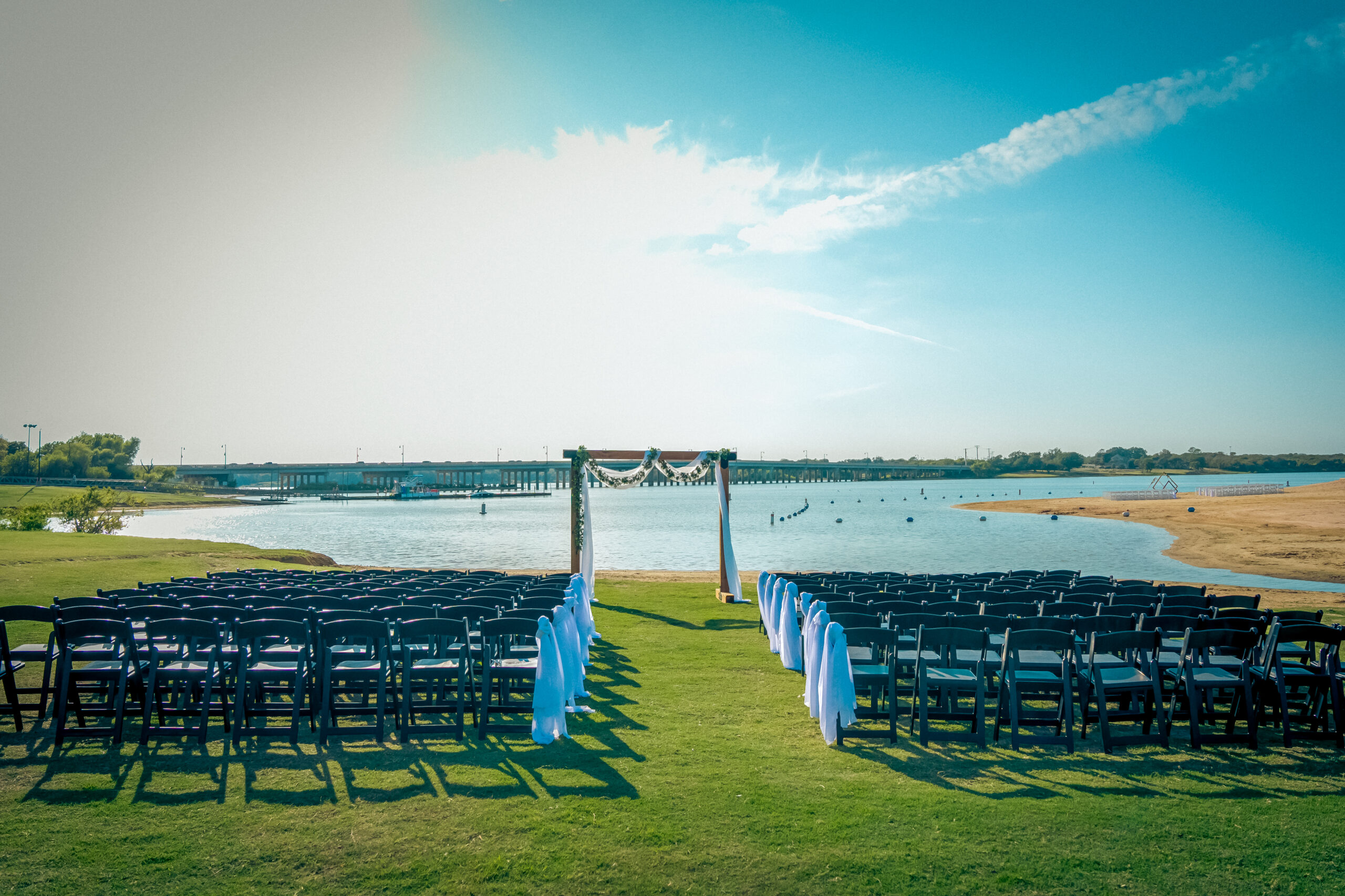 An enchanting outdoor wedding setup features rows of black chairs facing a flower-adorned archway on a lush grassy area, all framed by the stunning backdrop of the venues at the lakefront.