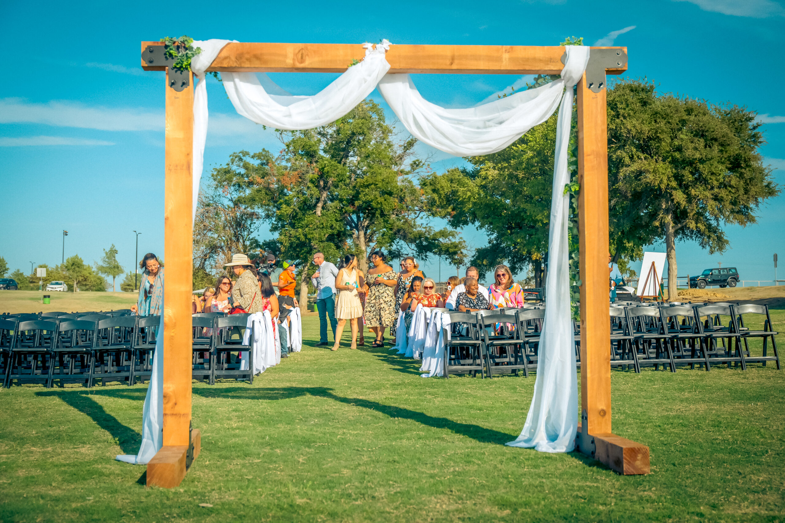 Outdoor wedding venue with wooden arch draped in white fabric, reminiscent of the serene settings at Lewisville Lake wedding venues. Guests are seated on black chairs under a clear blue sky, surrounded by lush trees and grass.