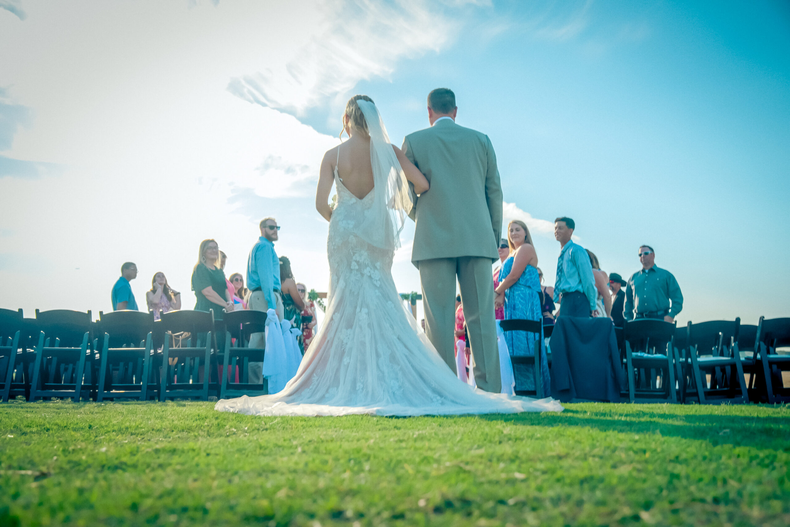 Bride and groom walking down an outdoor aisle at The Venues at the Lakefront, surrounded by guests under a clear blue sky. Their Lakeside wedding in Little Elm is as picturesque as Lewisville Lake itself.