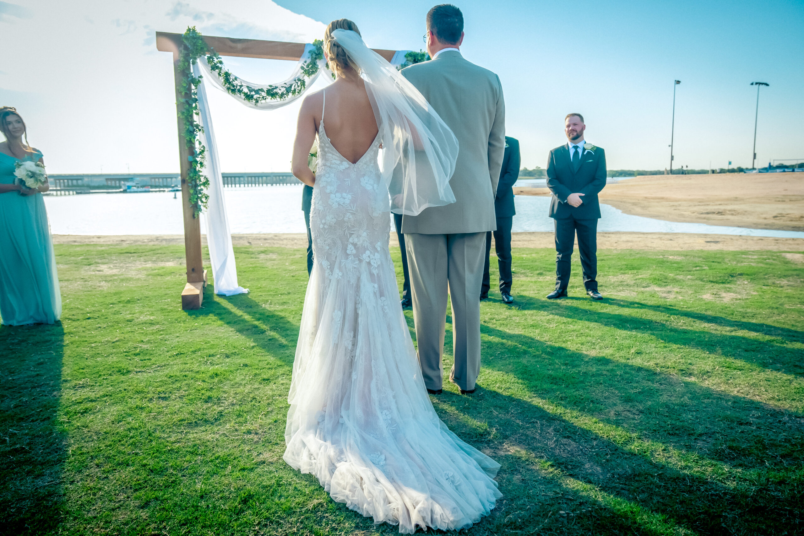 A bride and groom stand together at an outdoor wedding ceremony at one of the stunning Lewisville Lake wedding venues, beneath a wooden arch adorned with greenery. The scene overlooks a serene waterfront under a clear blue sky.
