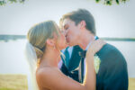 The bride and groom share a kiss outdoors on their lakeside wedding day in Little Elm. The bride wears a veil, and the groom dons a suit with a boutonniere, creating a picturesque moment at one of the charming North Texas wedding venues.