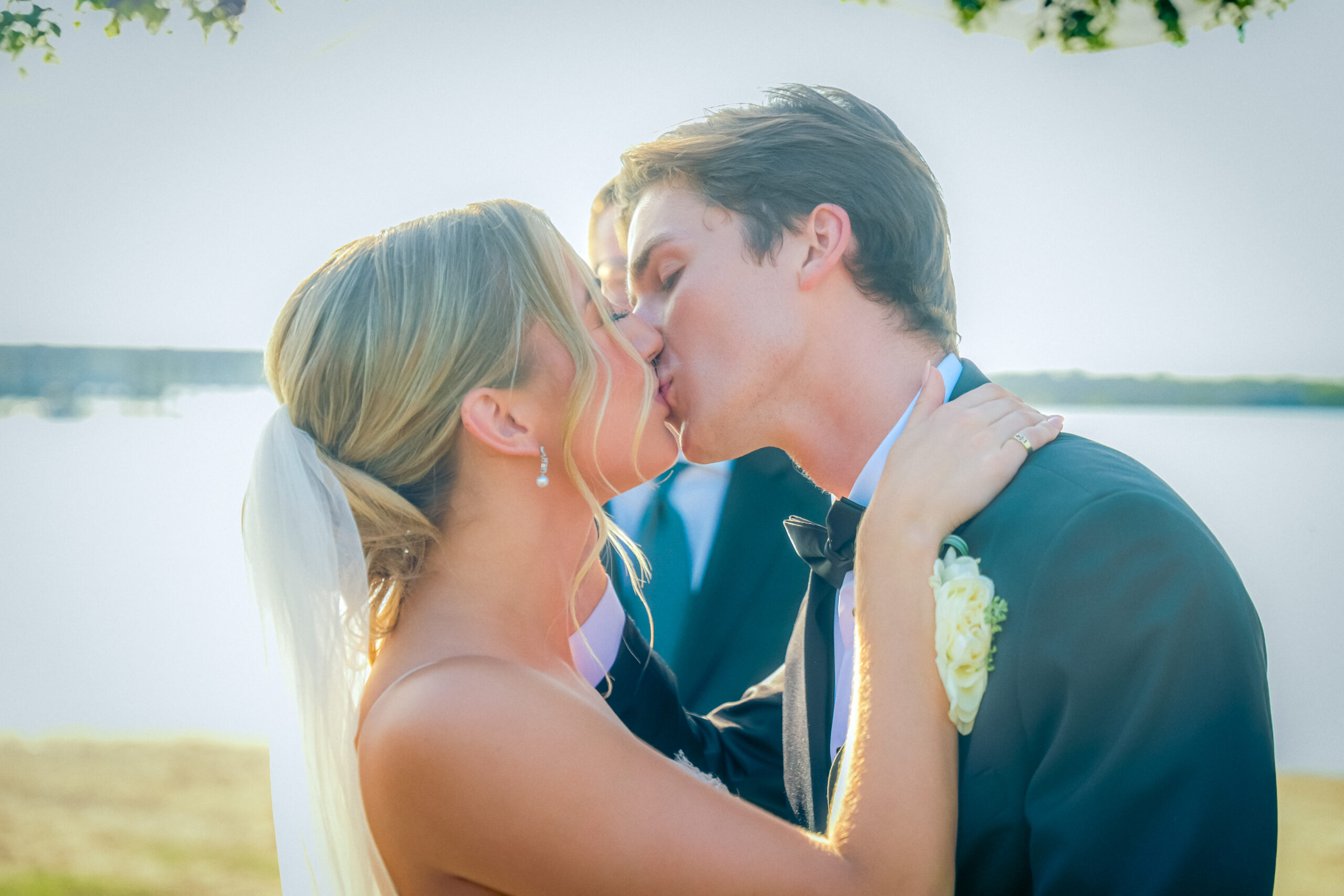 The bride and groom share a kiss outdoors on their lakeside wedding day in Little Elm. The bride wears a veil, and the groom dons a suit with a boutonniere, creating a picturesque moment at one of the charming North Texas wedding venues.