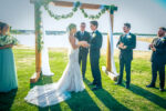 Bride and groom exchanging vows under a wooden arch adorned with greenery, by the serene Lewisville Lake, with bridesmaids and groomsmen standing nearby.