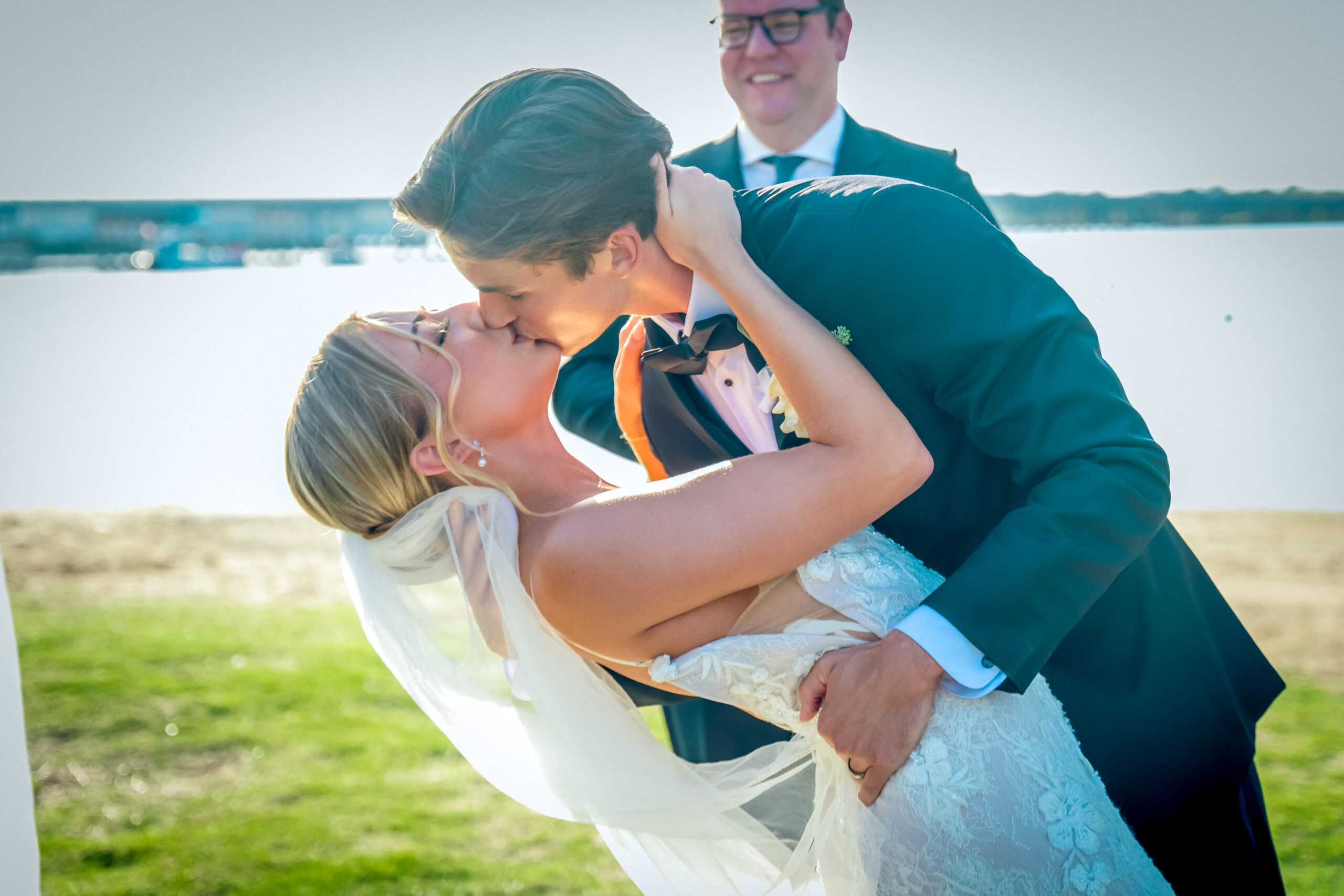 A newlywed couple kisses as the groom dips the bride backward outdoors at a picturesque lakeside wedding in Little Elm, with a smiling officiant in the background.