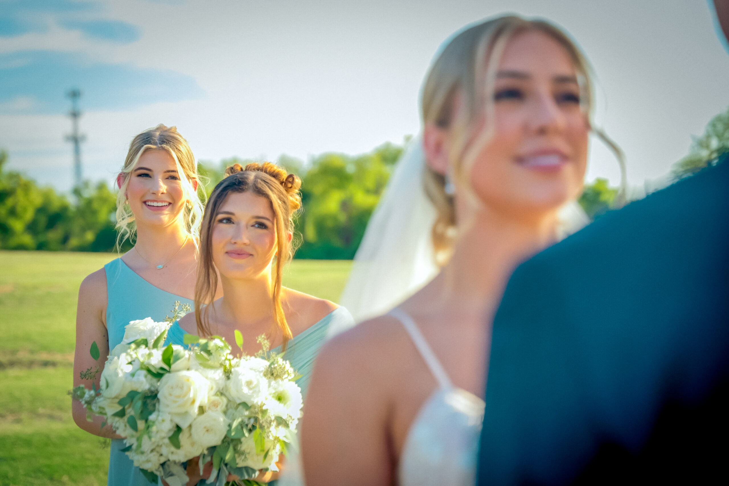 A bride and two bridesmaids in light dresses stand outdoors during a lakeside wedding in Little Elm. One bridesmaid holds a bouquet of white flowers as they enjoy the scenic backdrop of green trees and blue sky—a perfect day for those seeking beautiful Lewisville Lake wedding venues.