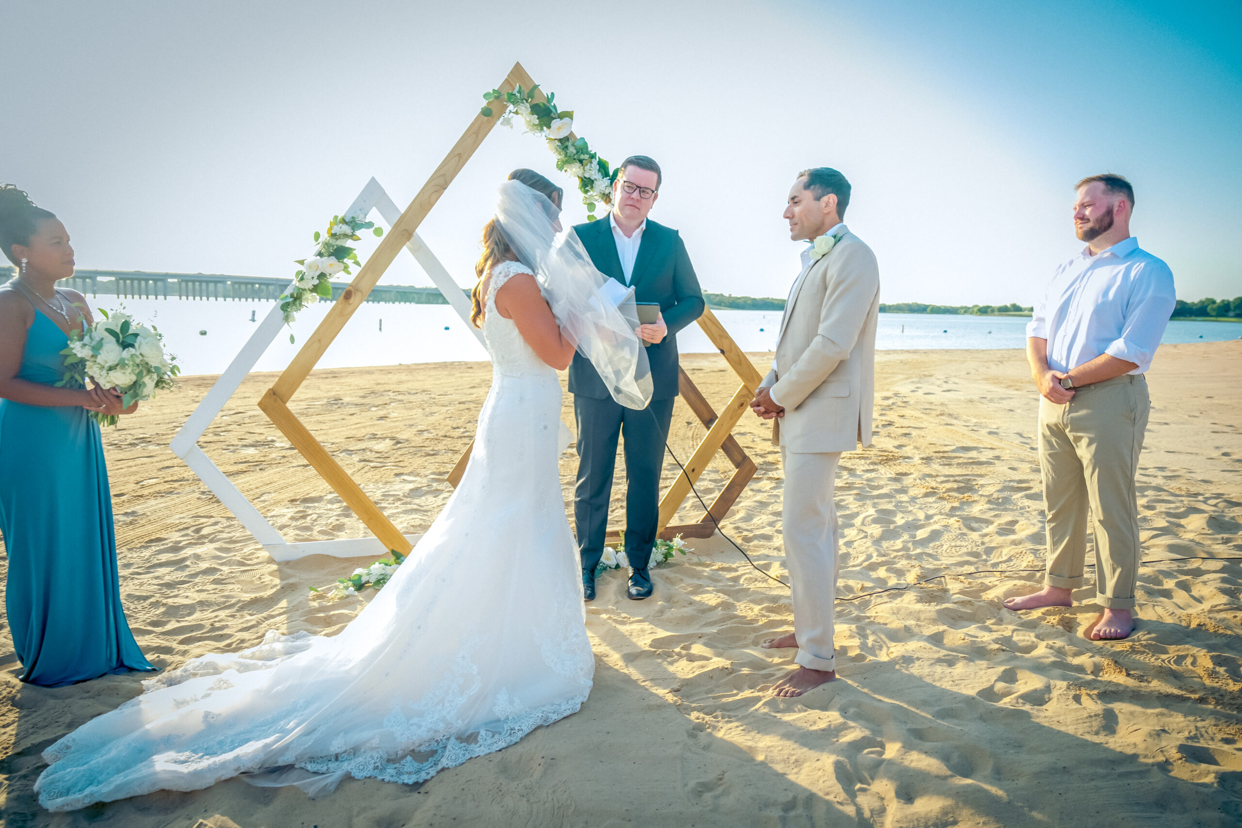 A couple exchanges vows on a sandy beach at a scenic North Texas wedding venue, with an officiant and two witnesses. Behind them stands a geometric arch decorated with greenery, adding charm to their intimate celebration.