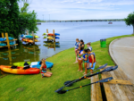 A group of children in life jackets listens to a woman near kayaks by a lake. Stacked kayaks and paddles are visible.