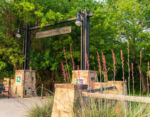 Entrance to Cottonwood Park Trail, marked by a stone and metal archway. Surrounded by green trees and red flowering plants.