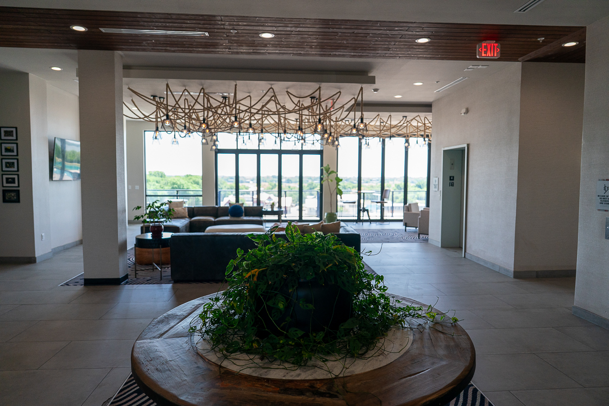 A modern lobby with a circular table topped with green plants in the foreground, seating area, large windows, and decorative light fixtures in the background.