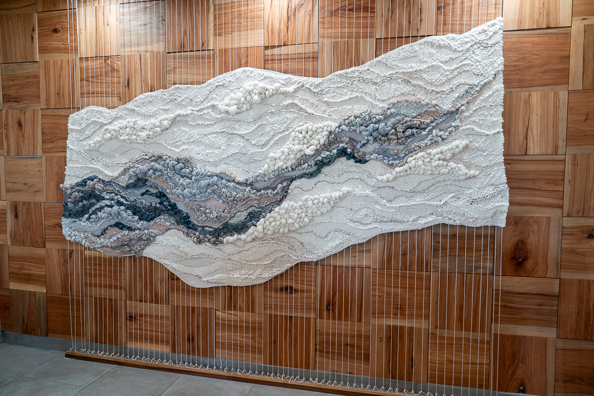 A large textured textile artwork with wavy patterns in white, gray, and blue hangs on a wooden wall. Thin vertical strings extend from the bottom of the piece to the floor.