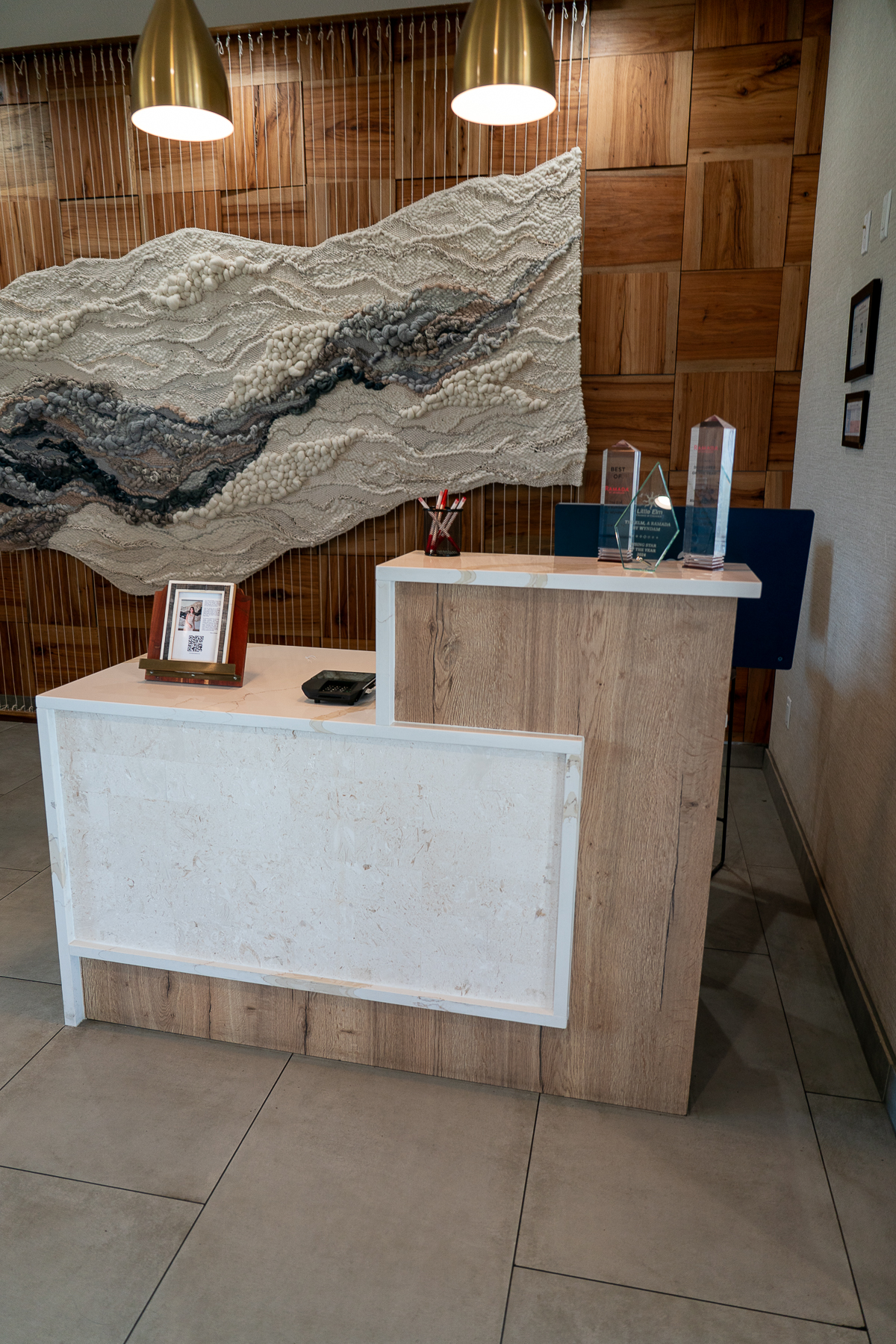 A modern reception desk with wood and white finishes, featuring awards, a framed photo, and office supplies, set against a decorative wall with textured art.