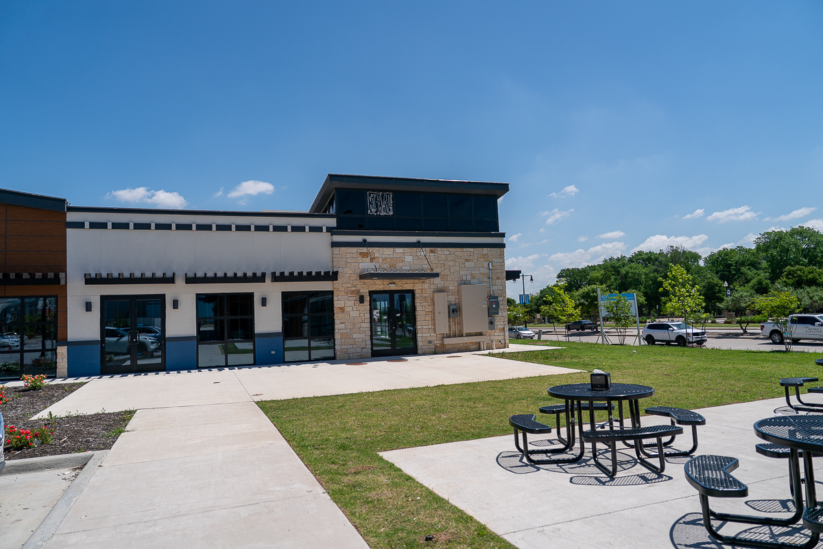 Outdoor seating area with round black metal tables and chairs in front of a modern building, under a clear blue sky.