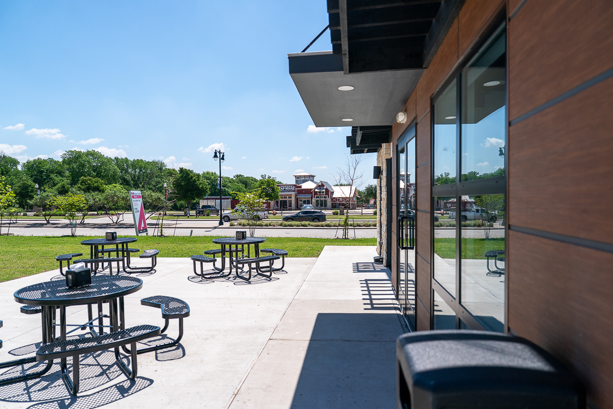 Outdoor patio area with black metal tables and benches on a sunny day, adjacent to a building with large windows, and a street with cars and buildings in the background.