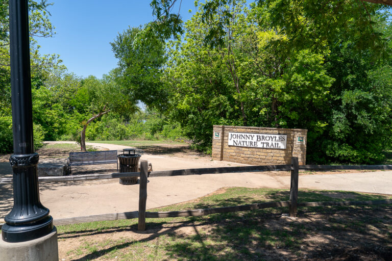Entrance to Johnny Broyles Nature Trail with a sign, bench, trash can, sidewalk, lamp post, and trees in the background.