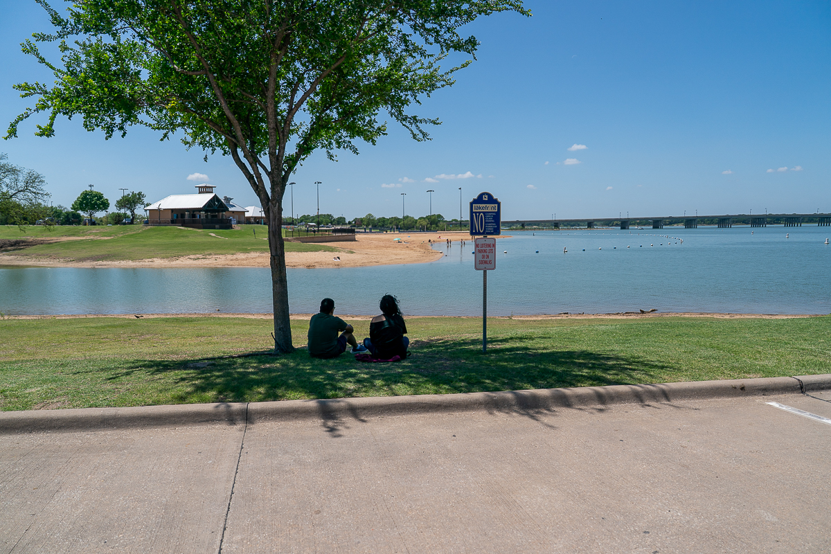 Two people sit under a tree by a lake, near a "No Parking" sign on the grass. A sandy beach, a building, and a bridge are visible in the background under a clear sky.
