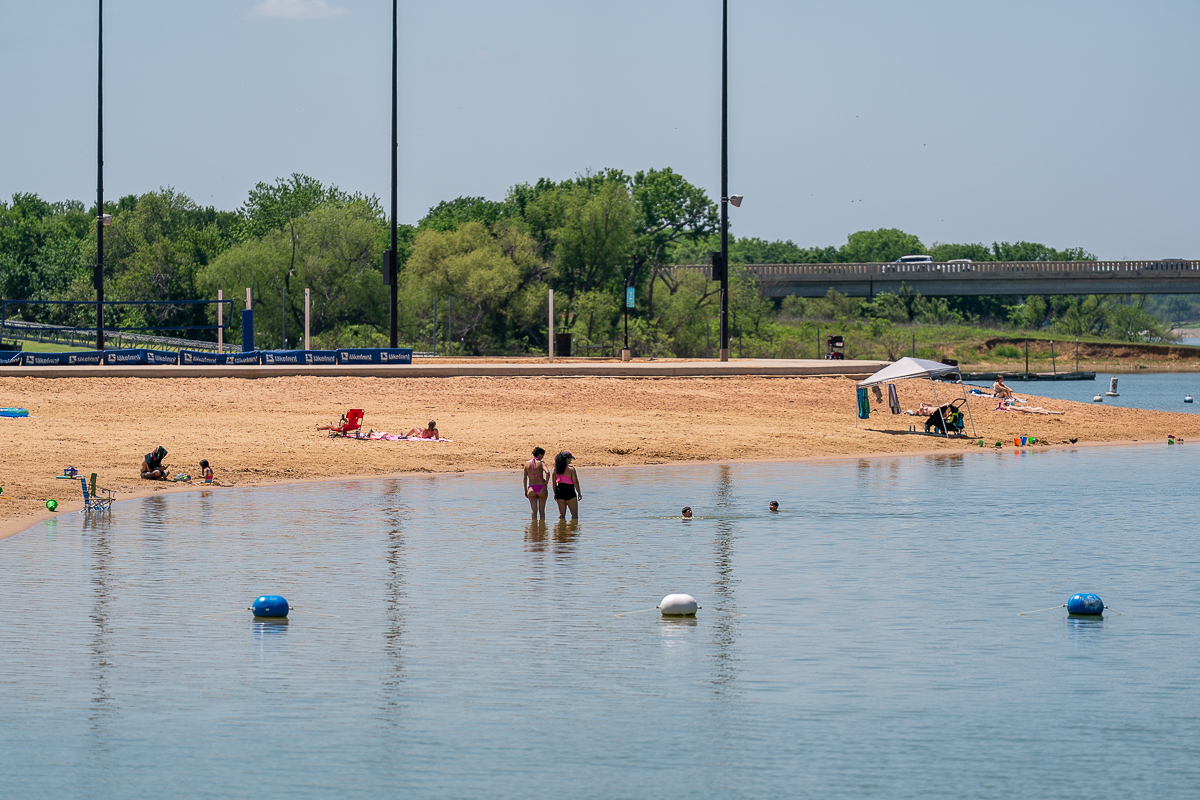 People relax and swim at a sandy beach beside a calm body of water, with trees and a bridge visible in the background.