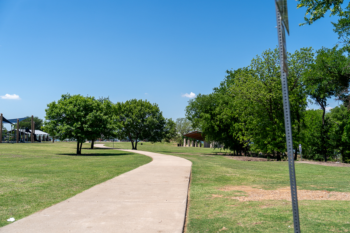 A paved path curves through a grassy park with scattered trees, a pavilion, and a metal signpost under a clear blue sky.