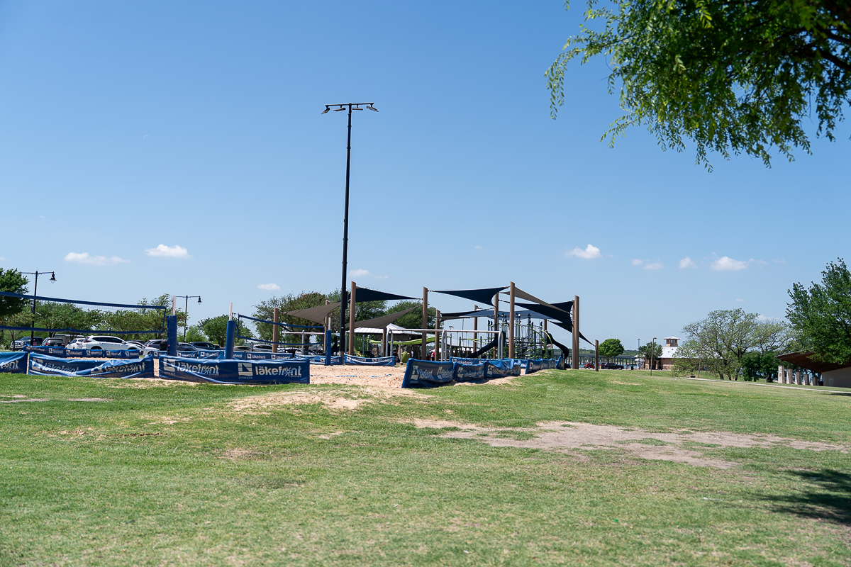 A public park with a sandy play area surrounded by blue fencing, shaded structures, green grass, and clear skies.