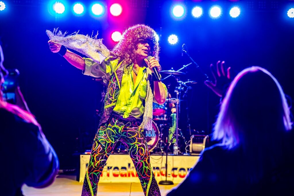 Performer in colorful attire sings energetically on stage, holding a microphone and silver jacket, with bright lights and audience visible.