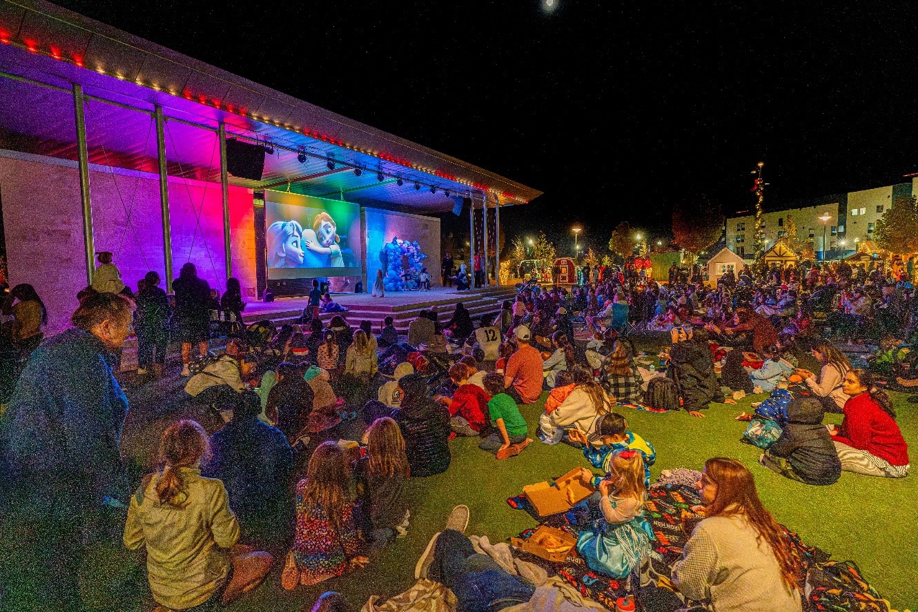 A crowd sits on a lawn at night, watching an animated movie on a large outdoor screen, with colorful stage lighting illuminating the scene.