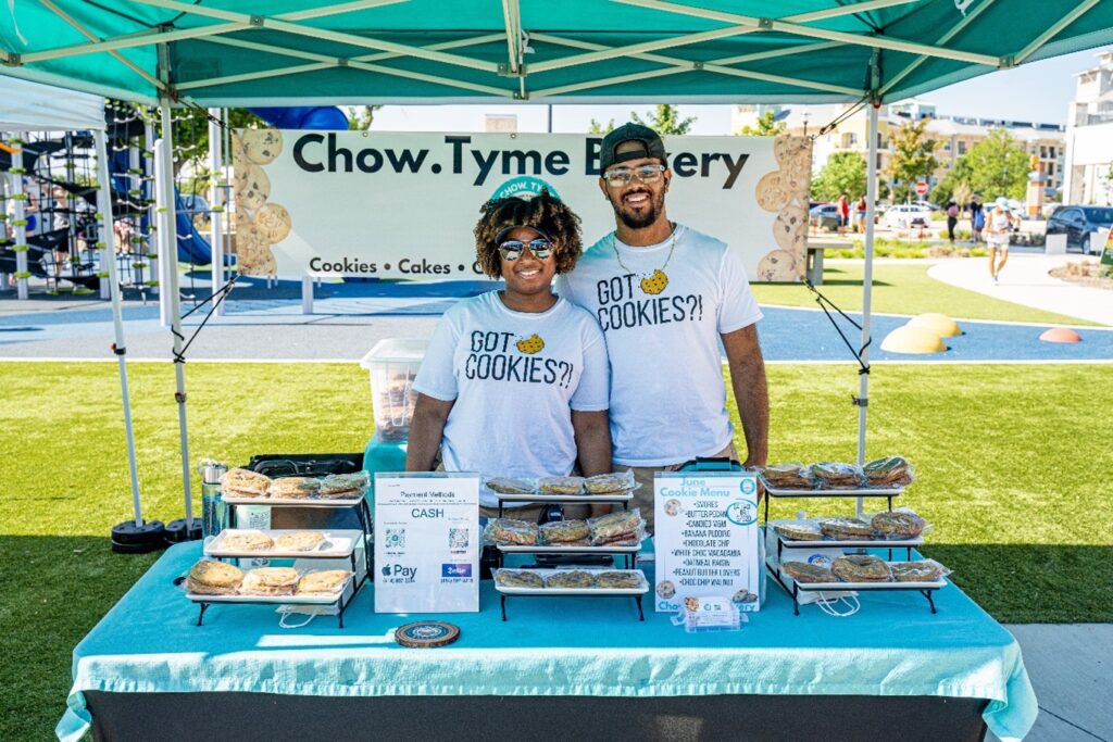 Two people stand behind a cookie stall under a green canopy at an outdoor market. The stall displays various cookies, a price list, and payment options. A banner reads "Chow.Tyme Bakery.