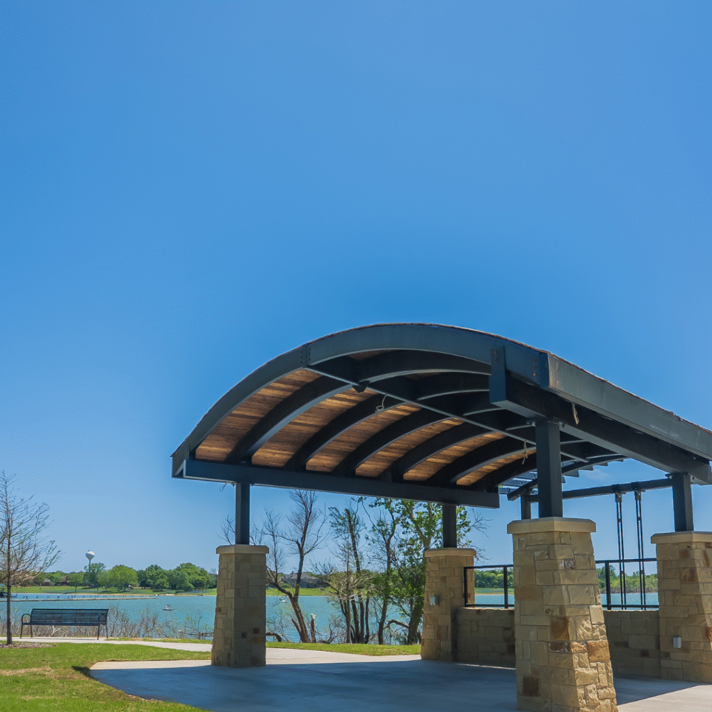 A modern outdoor pavilion with a curved roof stands near a river, perfect for lakeside weddings in Little Elm, surrounded by green trees under a clear blue sky.