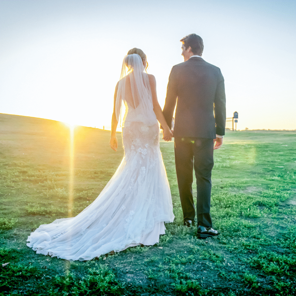 A bride and groom in formal attire walk hand in hand across a grassy field at sunset, capturing the romance of North Texas wedding venues.