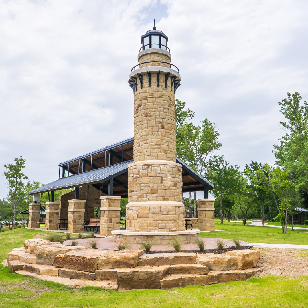 A tall, round stone lighthouse structure stands in front of a covered picnic pavilion in a park with green grass and trees, making it an ideal spot for lakeside weddings in Little Elm or those seeking unique North Texas wedding venues.