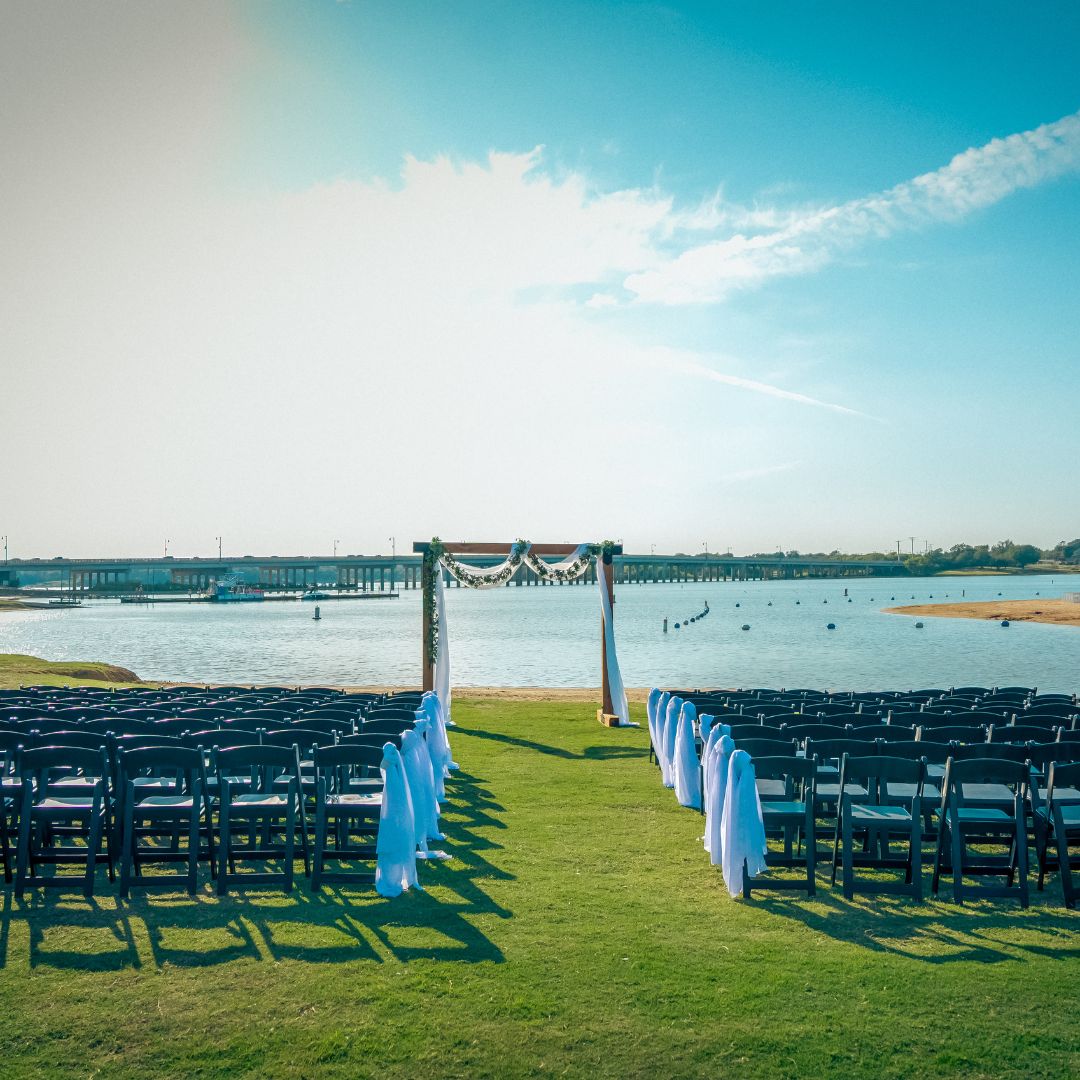 Rows of chairs face a beautifully decorated wedding arch set on grass by shimmering water and a clear blue sky—a perfect scene for beach weddings in Texas at a Marina wedding venue in Little Elm.