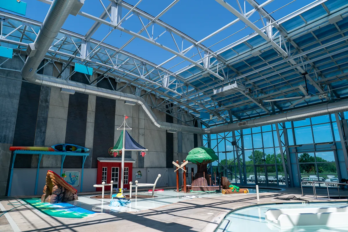 Indoor aquatic play area with colorful structures, a splash pad, and a shallow pool, The Cove at The Lakefront offers water fun under a high ceiling with large windows letting in natural light.